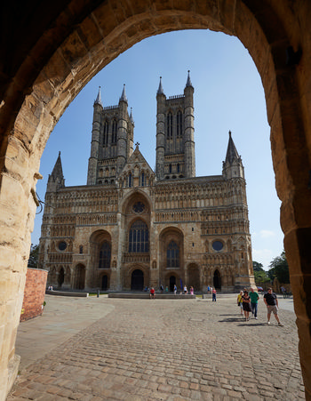 Lincoln cathedral arch This architecture photograph captures the impressive façade of Lincoln Cathedral in Lincoln, England, viewed through the archway of the surrounding stone structure during a late summer morning. The cathedral, a historic church and a prominent landmark in the United Kingdom, dominates the scene with its twin towers and intricate stone detailing under clear blue skies. The foreground shows people walking and standing on the expansive cobbled plaza, emphasizing the scale and grandeur of Lincoln Cathedral. This image highlights the architectural heritage of Lincoln and the importance of this cathedral church in the cultural landscape of England.